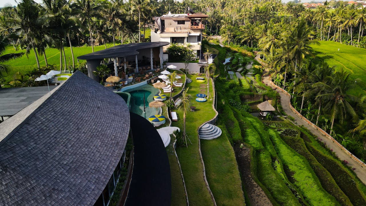 Stunning aerial shot of a luxury resort in Bali, Indonesia, surrounded by green rice terraces and palm trees under a sunny sky.